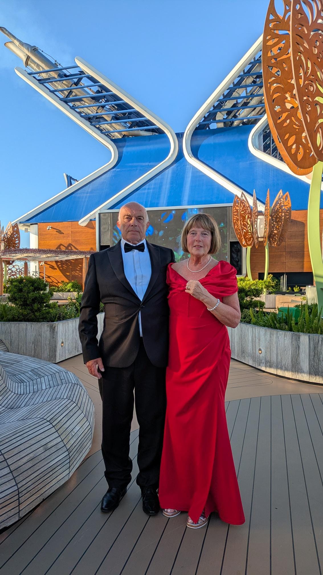 A man in a tuxedo and a woman in a red dress on the deck of Celebrity Apex, a cruise ship, with an 'X' shaped strucutre in the background
