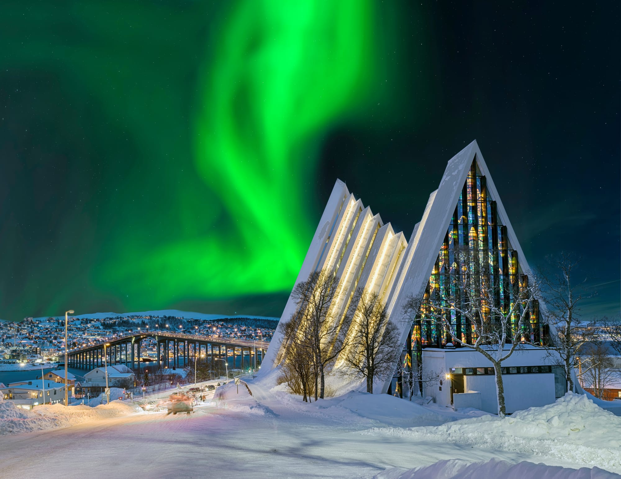 A modern cathedral in a snowy Norwegian town with a triangular facade and the northern lights' aurora borealis in the sky above